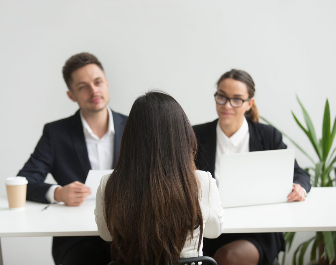 Back close up view of female applicant being interviewed by two HR managers reading her resume, checking data on laptop, asking questions for job position. Employment, hiring, first impression concept